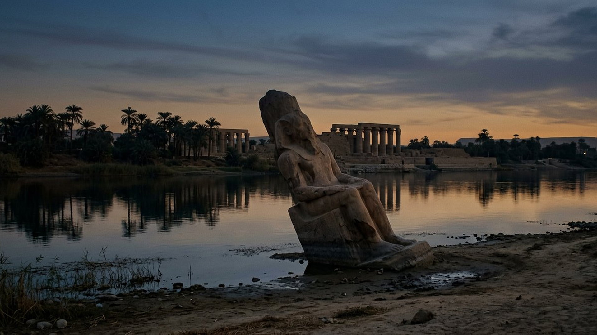 Statue colossale renversée au bord du Nil au crépuscule, temple en arrière-plan