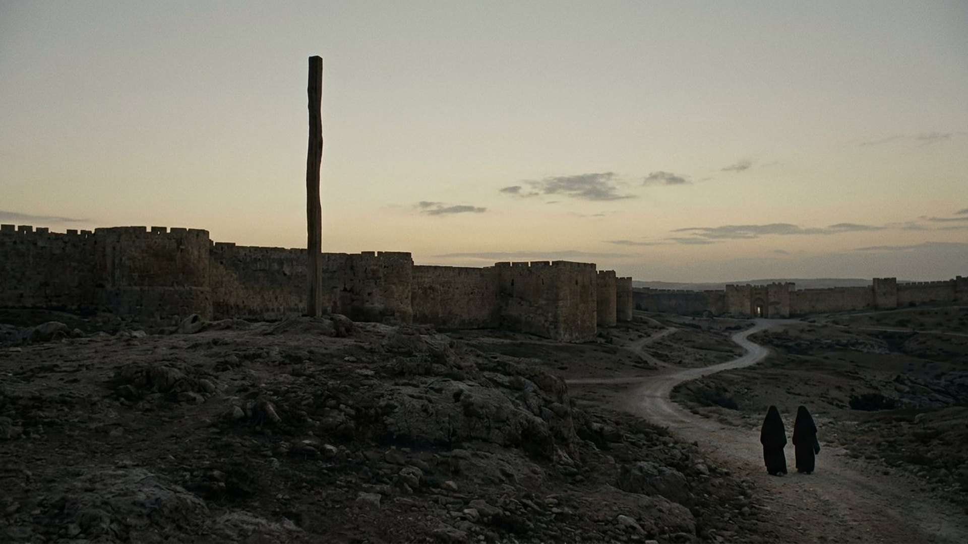 Un poteau vertical unique et vide dressé sur une colline à l'aube devant des murailles antiques, deux femmes en deuil au loin