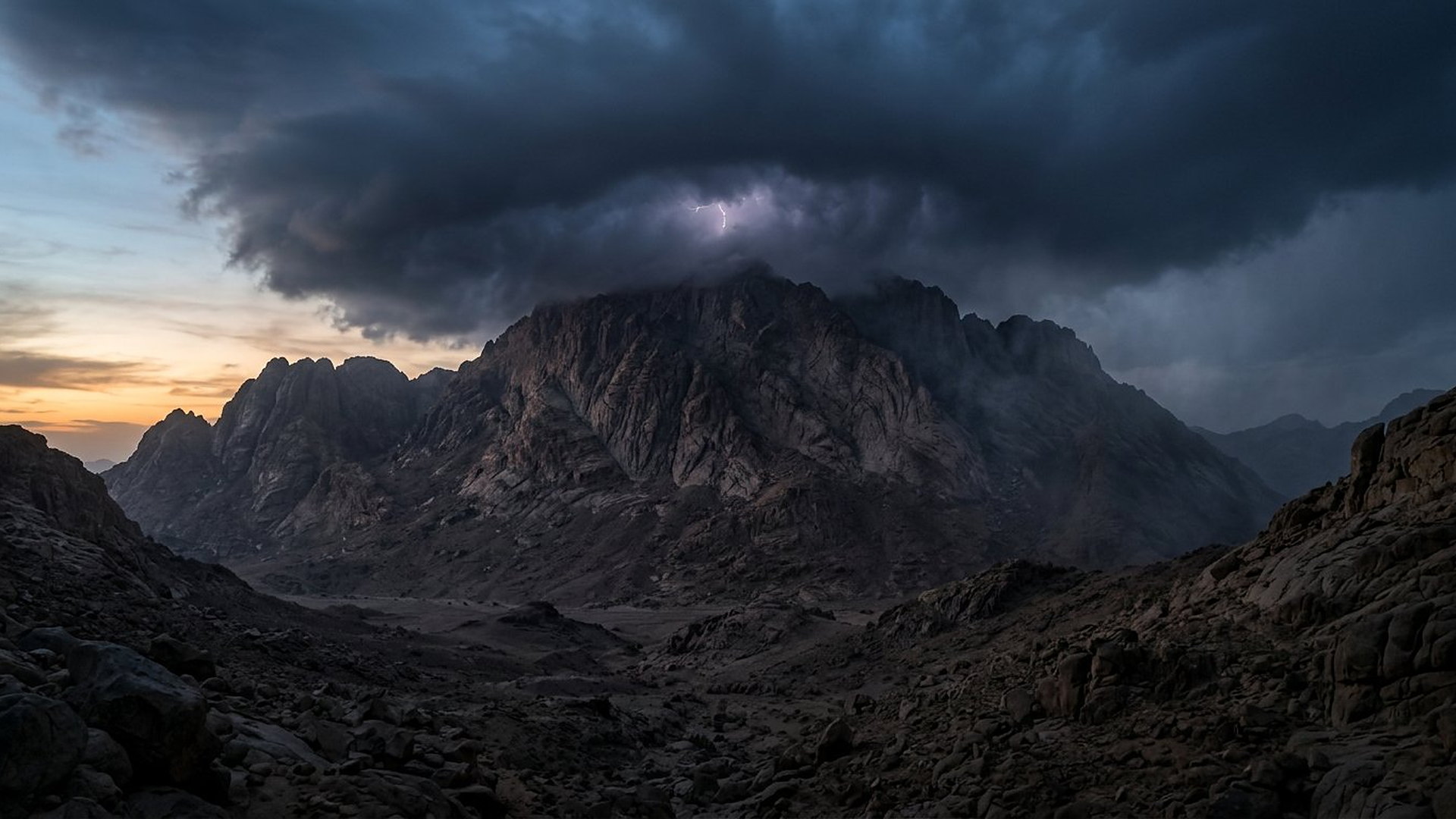 Massif de granit sombre sous un lourd nuage d’orage, un éclair discret dans la nuée, vallée rocheuse déserte au premier plan